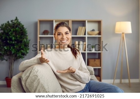 Smiling young woman blogger influencer sitting in front of webcam conducts online broadcast. Young woman sitting on sofa at home making video conference or recording video for blog. View webcam. Royalty-Free Stock Photo #2172691025