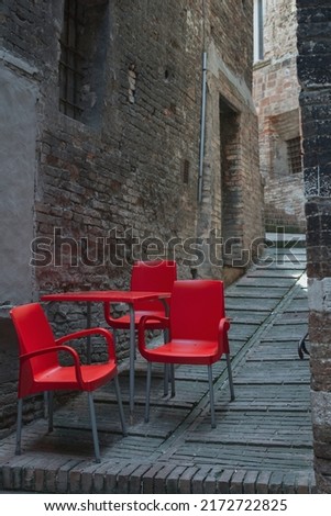 Three red chairs and a small table in an uphill lane. Small bars in an old Italian town. Pop up effect and color contrasts. Modern objects and medieval cities.	     