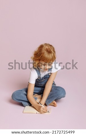 Pretty long-haired girl in a denim overalls sits cross-legged on a pink studio background. Makes notes in a notebook.