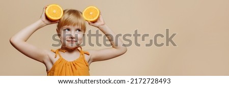 Banner photo of a cheerful little girl standing against an orange background. He holds half an orange in his hands and makes ears for himself.