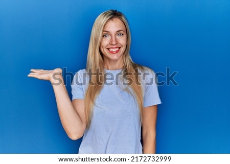 Beautiful blonde woman wearing casual t shirt over blue background smiling cheerful presenting and pointing with palm of hand looking at the camera. 