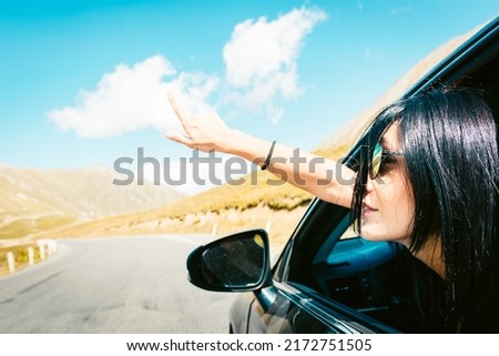 Female caucasian woman from car window points finger up left with asphalt road background. Copypaste road trip concept.