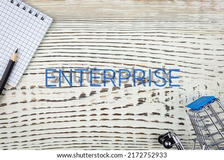 ENTERPRISE - word (text), on a white wooden table, a notebook and a trolley, a grocery basket. Business concept: buying, selling, commerce (copy space).