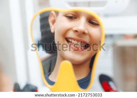 boy looks in the mirror with a toothy smile, sitting on a chair with a dentist in the dental office.