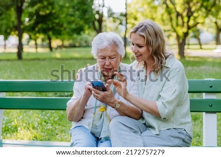 Adult granddaguhter helping her grandmother to use cellphone when sitting on bench in park in summer. Royalty-Free Stock Photo #2172757279