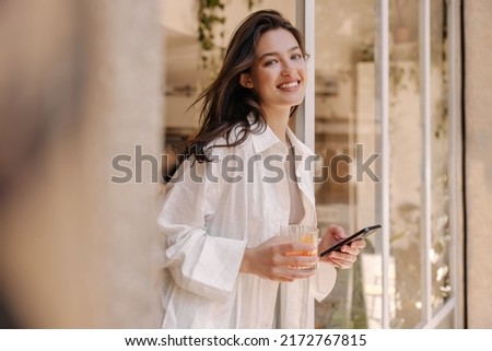 Beautiful young caucasian woman smiling looking at camera, holding phone and glass standing in doorway. Brunette wears white shirt warm weather. Mood, lifestyle, concept.