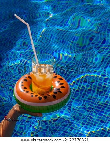 Cold refreshing drink with ice and a straw in the inflatable ring in shape of watermelon in a woman's hand in a pool of blue water. Concept of summer holiday in hotel or pool party. Copy space Royalty-Free Stock Photo #2172770321