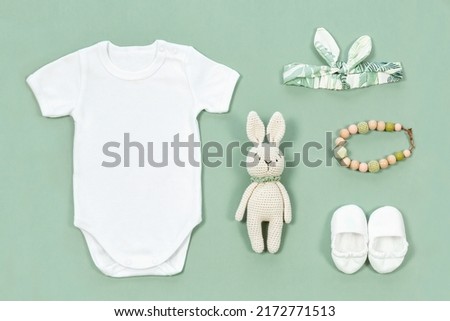 White bodysuit for baby mock-up. Mockup of infant clothes on a mint green background with baby toys and accessories. Crocketed toy of rabbit, cute head band, booties and teather beads next to a romper