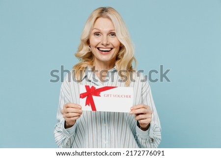 Elderly smiling caucasian woman 50s in striped shirt hold gift certificate coupon voucher card for store isolated on plain pastel light blue color background studio portrait. People lifestyle concept