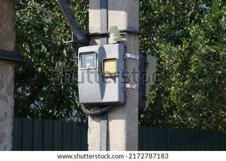 one gray electric meter hangs on a concrete pole in the street against the backdrop of a fence and green vegetation