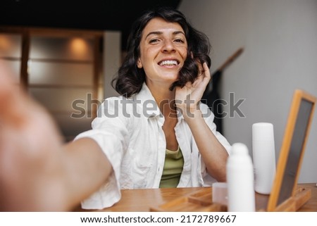 Smiling young caucasian woman with snow-white smile makes photo sitting in room. Brunette with bob haircut is wearing shirt. Lifestyle and beauty concept