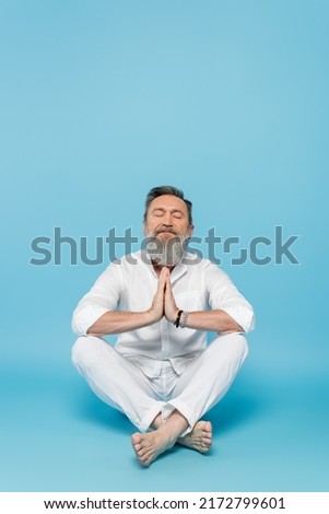 bearded man with closed eyes and praying hands meditating in easy pose on blue