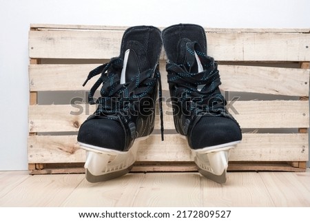 A pair of black hockey skates with well-sharpened metal blades in a white plastic frame designed to play ice hockey as a popular winter sport stay next to the craft wooden wall waiting for game starts