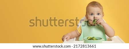Cute little baby wearing bib while eating on yellow background, space for text. Banner design Royalty-Free Stock Photo #2172813661