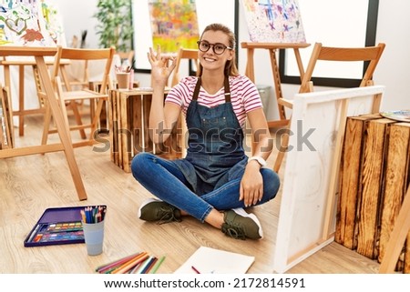 Young brunette woman at art studio sitting on the floor smiling positive doing ok sign with hand and fingers. successful expression. 