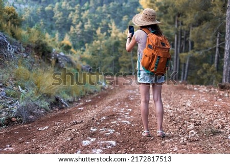 A girl photographs a beautiful mountain landscape on a smartphone, a woman in a hat and with a backpack walks along a forest path and takes a selfie