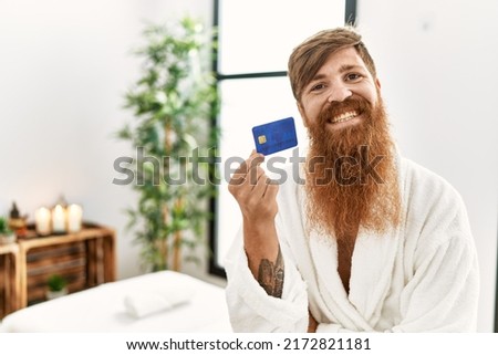 Young redhead man wearing bathrobe holding credit card at beauty center