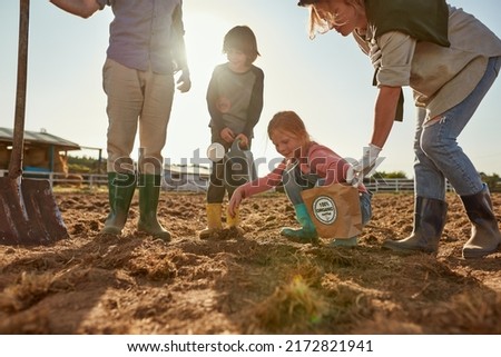 Mother, father and son waiting daughter throwing organic seeds in ground hole on plow field on ranch or farm. Young family spend time together. Eco harvest. Modern countryside lifestyle. Agriculture Royalty-Free Stock Photo #2172821941