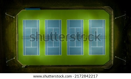 Evening aerial photo of outdoor blue tennis courts with pickleball lines and green out of bounds area with lights turned on.	