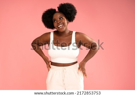 Attractive female model with dark skin and afro hairstyle posing on pink pastel studio background, smiling and showing her belly. Diet concept. Body positive, conscious. Real people emotions.