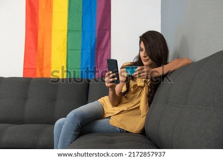 young adult woman sitting on the couch shopping online with her smartphone with a rainbow flag in the background