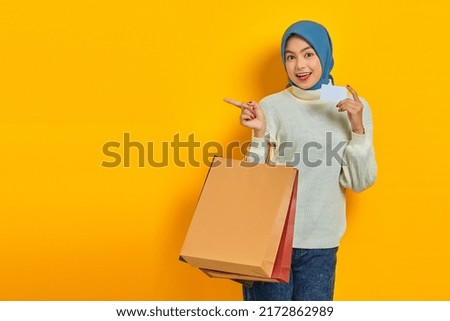 Cheerful beautiful Asian woman in white sweater holding shopping bag and credit card, pointing finger aside isolated over yellow background
