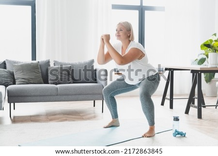 Plus size plump caucasian young woman athlete doing home workout training, losing weight, doing squats in fitness sporty clothes, slimmimg indoors Royalty-Free Stock Photo #2172863845