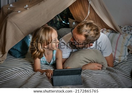 Portrait of a 6 year old boy and his father having fun playing in teepee tent. Father and son using digital tablet watching cartoons or playing computer games lying in kid tent at home. 