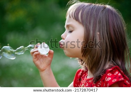 Little child girl in red dress blowing soap bubbles on a summer day at green nature background. Childhood and playful concept.