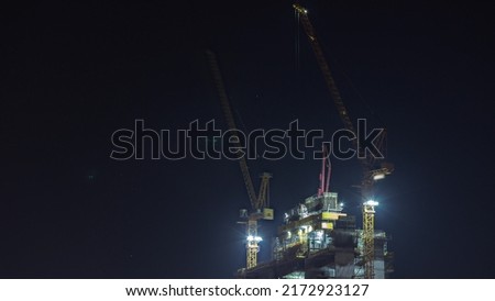 Skyscraper under construction with cranes night timelapse. Building of new multi-storey tall tower with builders working on top