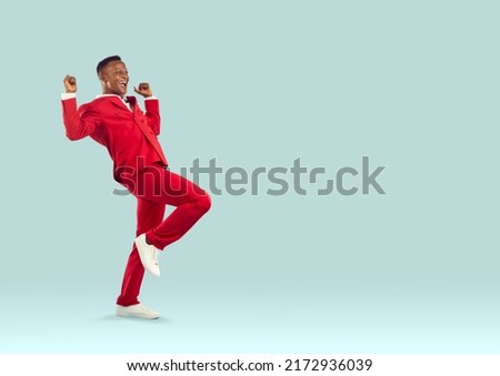Happy cheerful confident attractive Afro American man in cool modern funky red suit celebrating victory, feeling overjoyed and super excited, dancing and having fun on light blue copyspace background