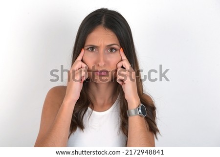 Serious concentrated young beautiful caucasian woman wearing white top over white background keeps fingers on temples, tries to ease tension, gather with thoughts and remember important information.