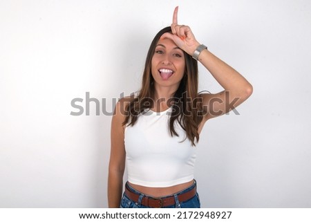 young beautiful caucasian woman wearing white top over white background gestures with finger on forehead makes loser gesture makes fun of people shows tongue