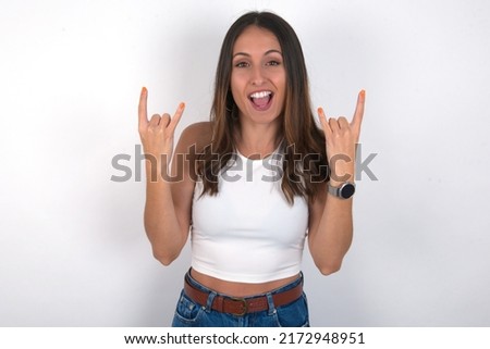 young beautiful caucasian woman wearing white top over white background makes rock n roll sign looks self confident and cheerful enjoys cool music at party. Body language concept.