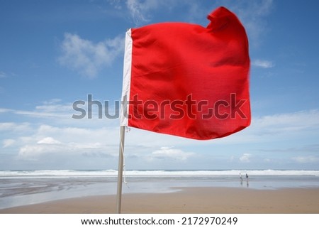 Red flag on the Beach. A red flag flies in the wind on a windy but sunny summer's day, warning swimmers of dangerous surf conditions. Red flag, Cornish Beach, summer, sand, blue sky, white water  Royalty-Free Stock Photo #2172970249