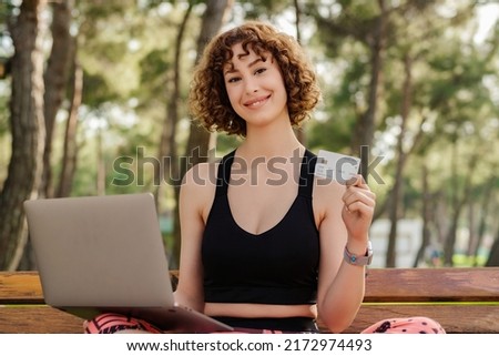 Cute redhead woman wearing sports bra standing on city park, outdoors using laptop computer and holding credit card. Online shopping, banking and transaction concepts.