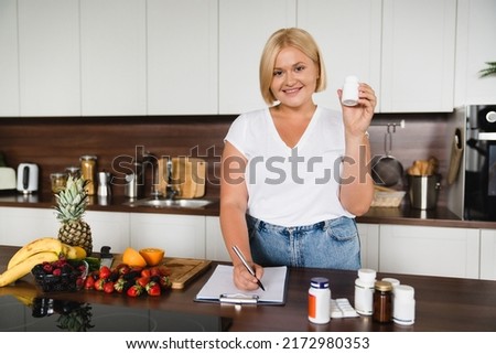Caucasian young plus size plump woman nutritionist writing counting calories healthy food supplements for slimming shaping dieting, holding pills medicines looking at camera at home kitchen