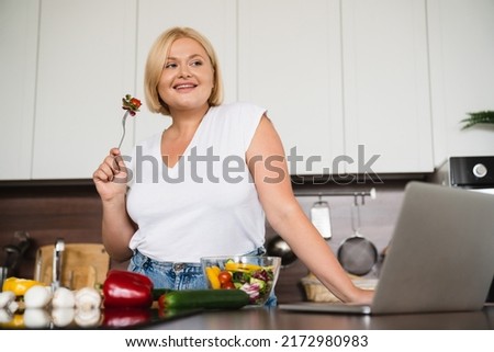 Low angle view plus size plump woman culinary vlogger blogger eating vegetable salad, preparing healthy vegan vegetarian food using laptop for blogging online at home kitchen