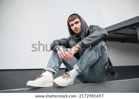 Fashionable handsome young hipster man wearing trendy clothes with jacket, hoodie, jeans and sneakers sitting near the white wall on the street Royalty-Free Stock Photo #2172985697