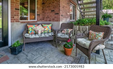 Panorama Woven couch and armchairs on an outdoor patio under the deck of a house with bricks. There is a glass front door on the right side near the windows and a view of a stairs on the left.