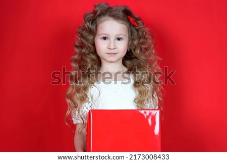Portrait of happy positive cheerful small little kid child girl in dress isolated on red background holding red present big box, smiling, celebrating. Present, holiday, happiness concept 
