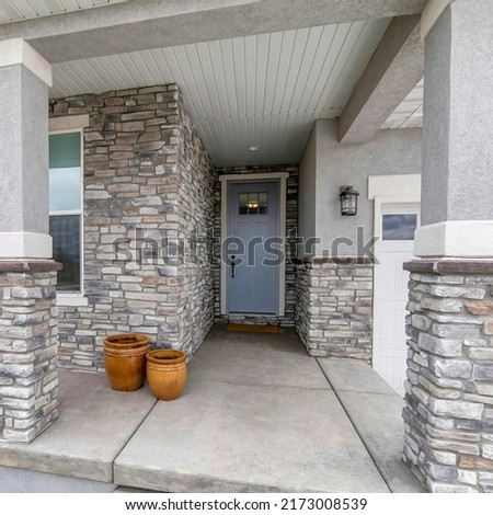 Square House exterior with stone veneer siding and garage. There are two empty pots near the front yard garden with large rock and window beside the gray front door near the white garage door. Royalty-Free Stock Photo #2173008539
