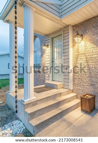 Vertical Exterior of a house with stone veneer siding and sectional garage door. There is an elevated light green front door with two wall lamps on the side and rain chain at the front. Royalty-Free Stock Photo #2173008541