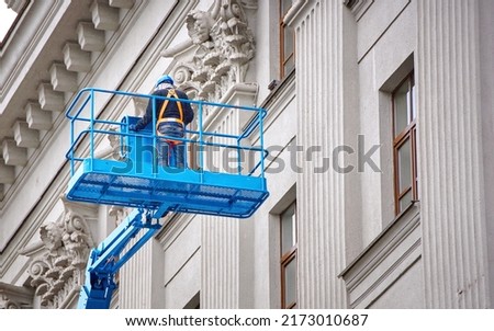 Construction worker in lift bucket of crane restore and repair historic facade of building. Man in cradle, restoring plaster decoration on facade. Workers painting building, renovating and repair work Royalty-Free Stock Photo #2173010687
