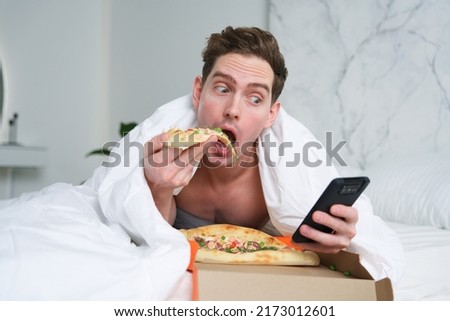 Funny hungry handsome young man is lying on bed in bedroom with blanket eating pizza and looking, using smartphone cell mobile phone. Junk fast food, hunger. Lazy weekend day. Delivery concept. Royalty-Free Stock Photo #2173012601