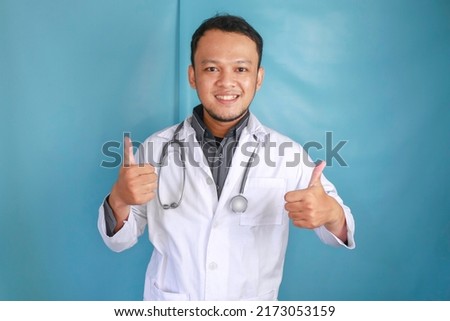 Portrait of a young Asian man doctor, a medical professional is smiling and showing thumbs up or OK sign isolated over blue background