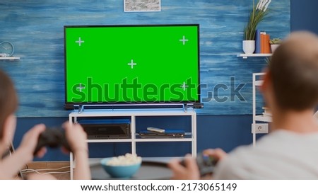 Closeup of couple holding controllers playing action console games on green screen tv sitting on couch and eating popcorn. Young gamers spending free time gaming online on chroma key mockup display.
