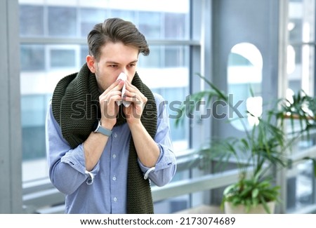 Portrait of sick ill handsome guy in scarf blowing his nose in paper handkerchief indoors at office building, young man with runny nose, flu. Symptoms of coronavirus, covid-19. Allergy, feeling unwell Royalty-Free Stock Photo #2173074689