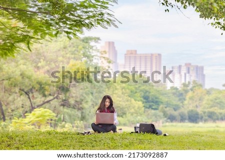 Asian woman relax with laptop outdoors. Smiling woman sitting on the grass with laptop, surfing the net and coffee in the morning before going to work at park on sunny day with city view on background