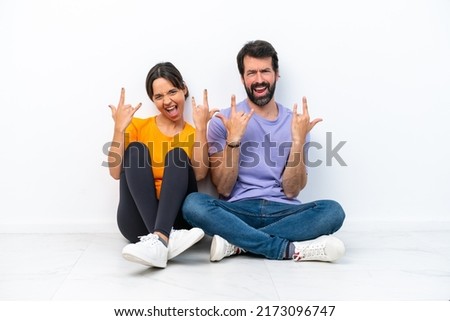Young caucasian couple sitting on the floor isolated on white background making rock gesture